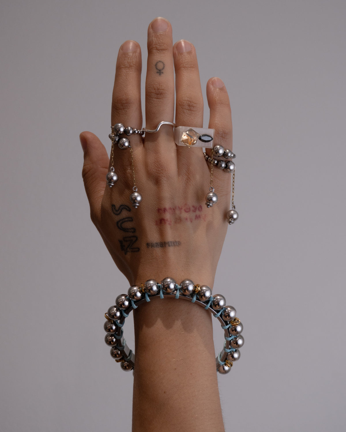 Collier bracelet en acier inoxydable. Il s'agit d'un collier composé d'un cordon en cuir brun avec un pendentif jonc argenté orné de perles argentées et de grains de café dorés tressés porté en bracelet jonc