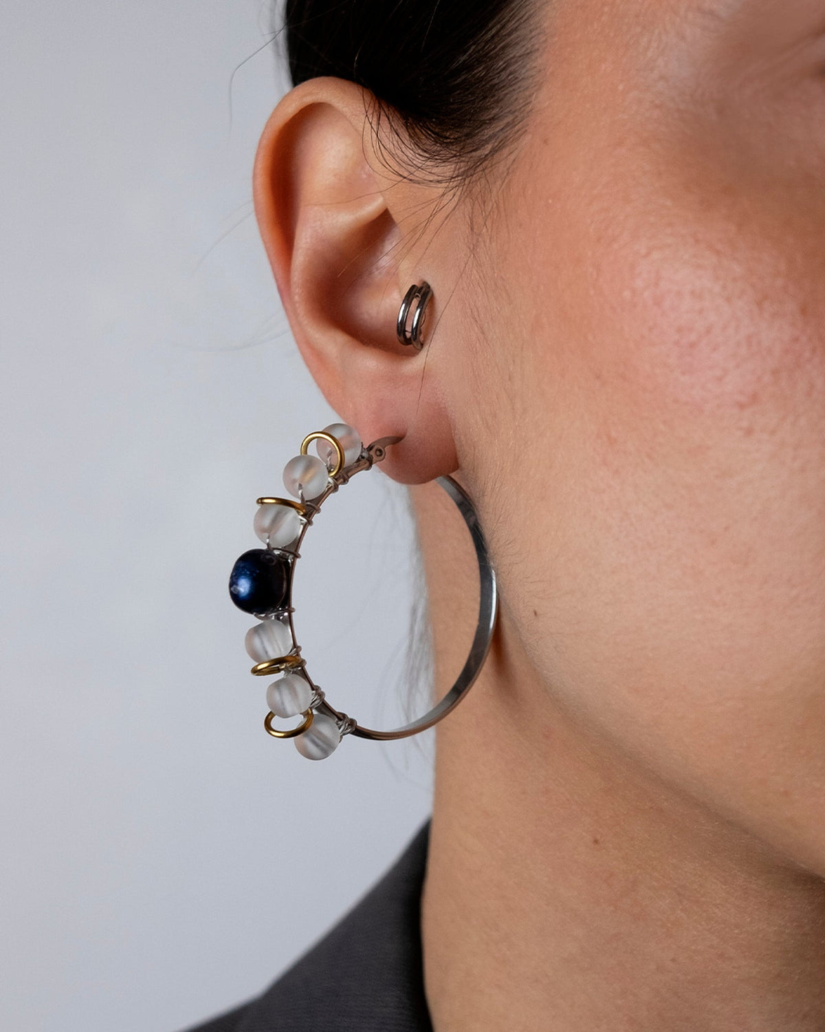 Mono boucle d'oreille en acier inoxydable porté composée d'une grande créole argentée et de différentes perles tressées en résine, d'une perle de culture d'eau douce noire et d'anneaux dorés
