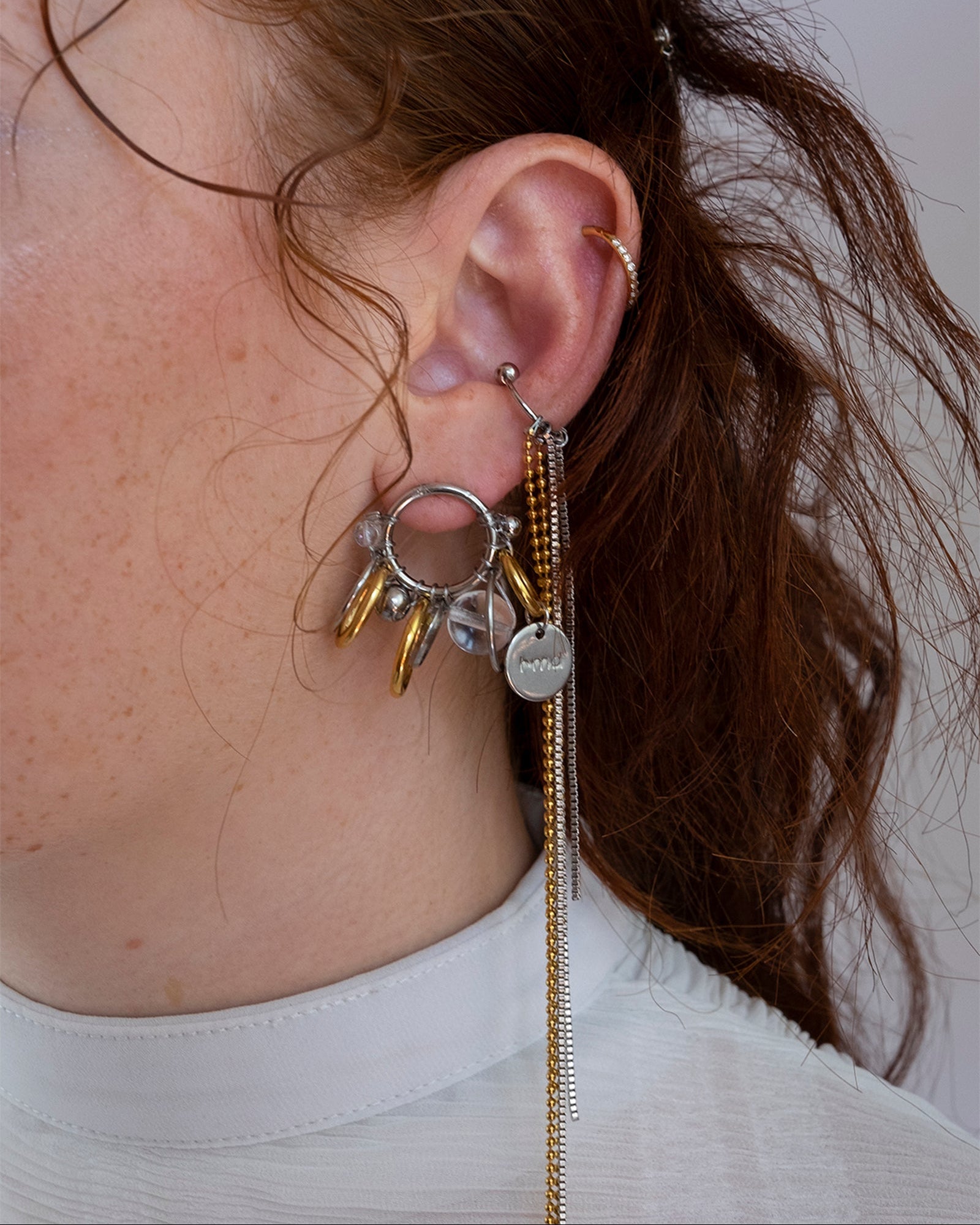 Mono boucle d’oreilles en acier inoxydable composée d’une mono boucle d'oreilles puce argenté, autour de laquelle sont tressés différents anneaux doré et argenté, des perles en verre transparentes et un médaillon de la marque