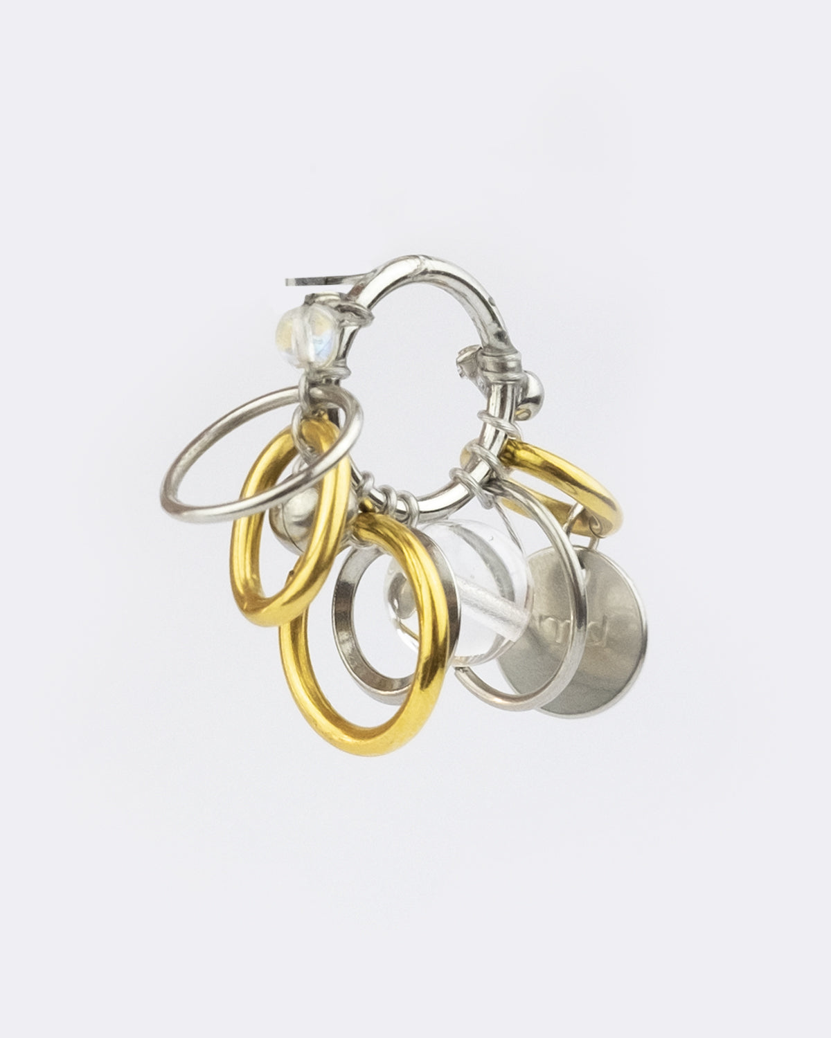 Mono boucle d’oreilles en acier inoxydable composée d’une mono boucle d'oreilles puce argenté, autour de laquelle sont tressés différents anneaux doré et argenté, des perles en verre transparentes et un médaillon de la marque