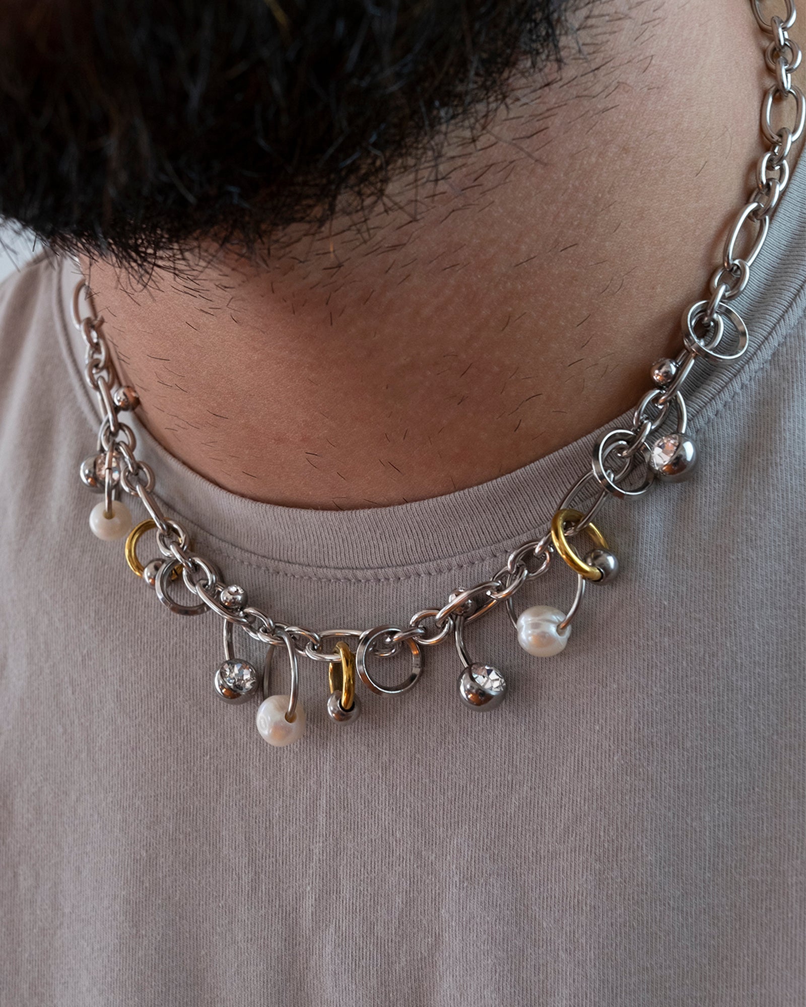 Collier en acier inoxydable. Il s'agit d'un collier composé d'une chaine argenté agrémenté d'anneaux de différentes tailles, de perles de culture d'eau douce ainsi que de piercing