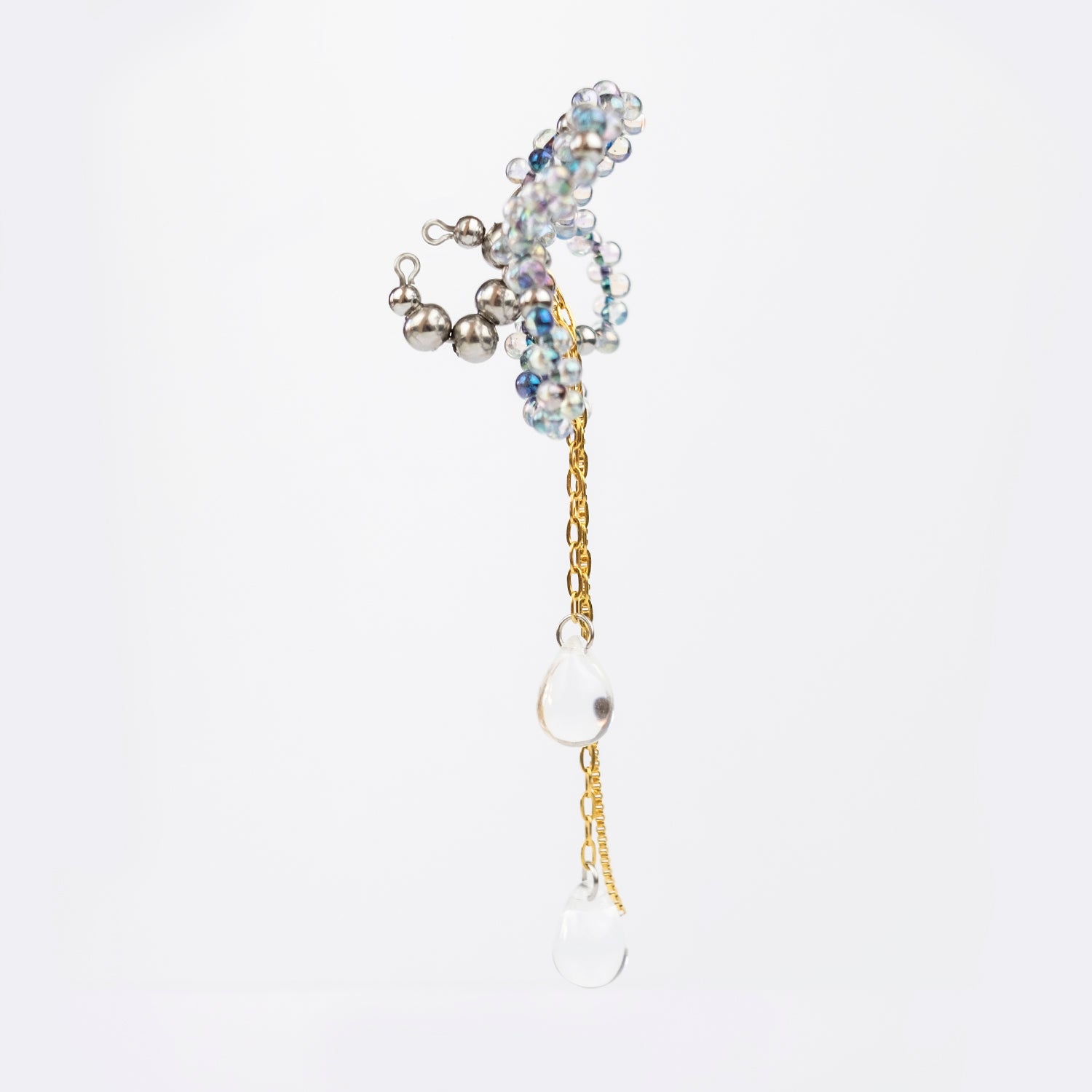 Earcuff en acier inoxydable. Il s’agit d’un faux piercing pour oreille non perçée qui se positionne au niveau du conch ou du lobe suivant la largeur de l’oreille. Il est composé d’un anneau argenté, d'une sculpture en perles en verre en forme de fleur et de chaine doré avec des perles goutte au bout