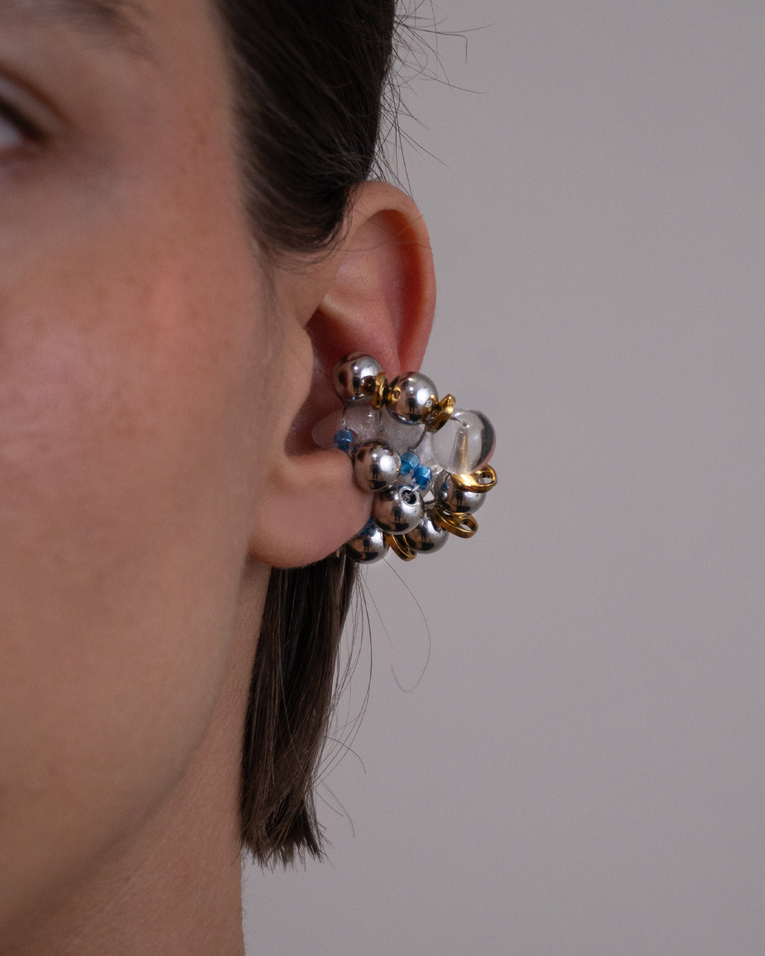 Earcuff Jelly Duo Bleu porté en plastique biosourcé composé d'une multitude de perles argentées de grains de café dorés et de perles en verre colorées bleues tressés sur une base transparente