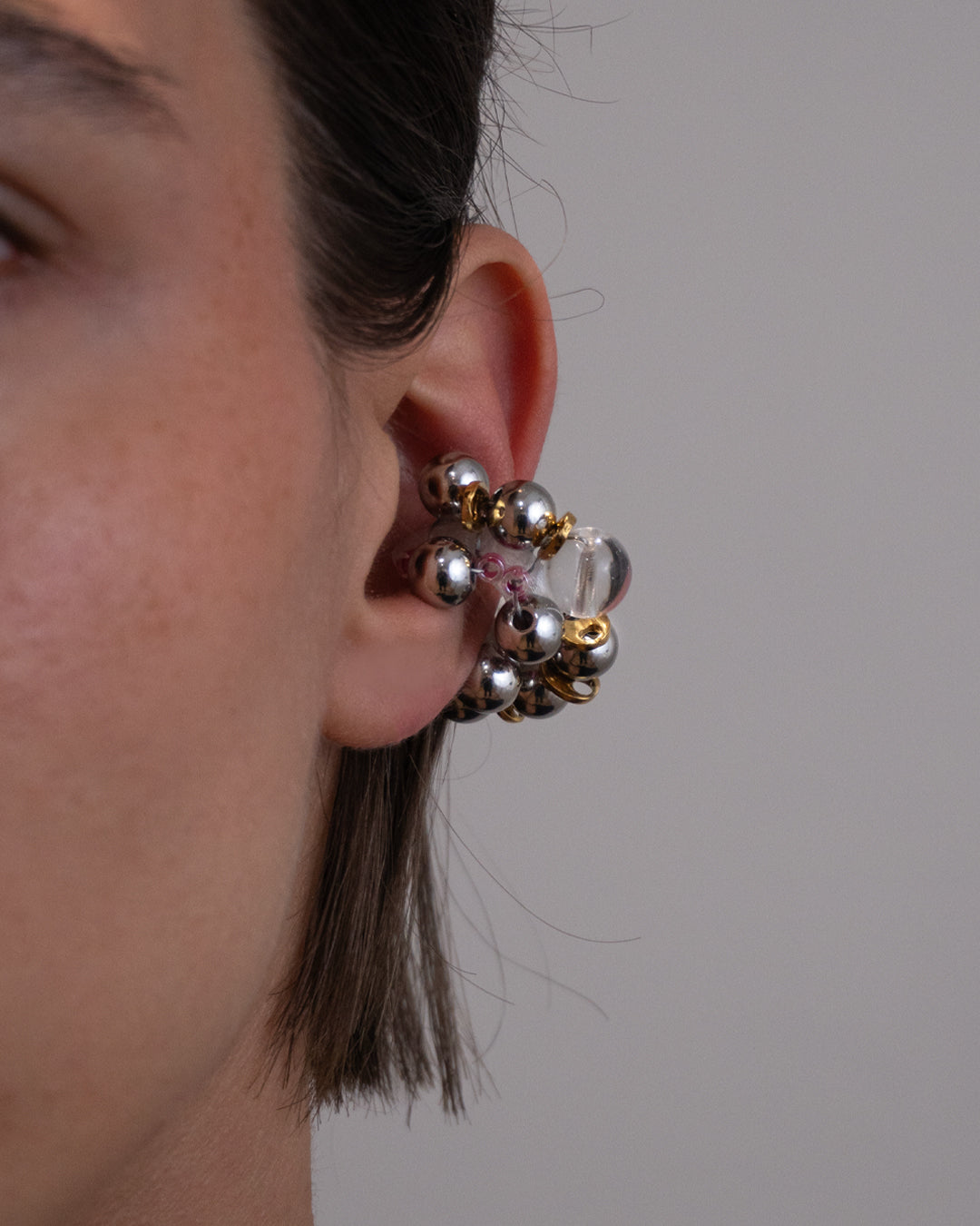 Earcuff Jelly Duo Rose porté en plastique biosourcé composé d'une multitude de perles argentées de grains de café dorés et de perles en verre colorées roses tressés sur une base transparente