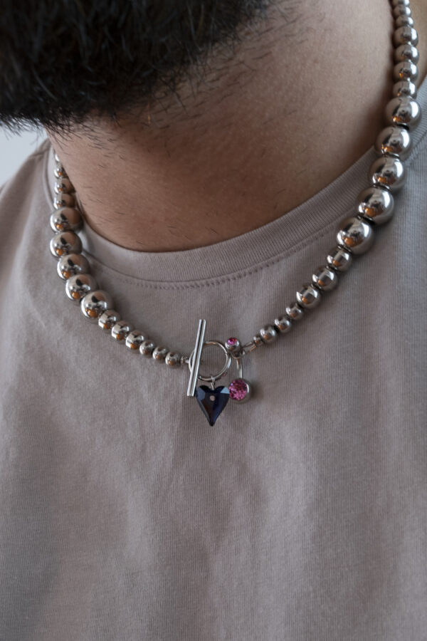 Collier en acier inoxydable. Il s'agit d'un collier composé de perles argenté de différentes tailles, d'un coeur en cristal et d'un piercing